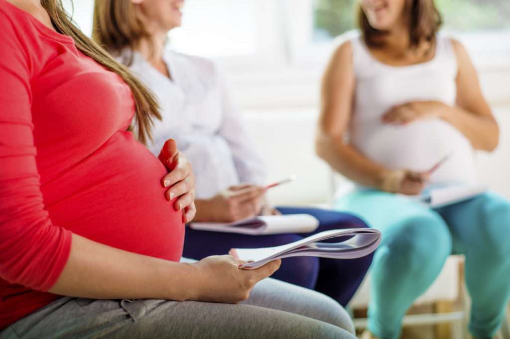 an image of a group of pregnant women sitting in a circle of chairs together and going over paperwork and chatting together